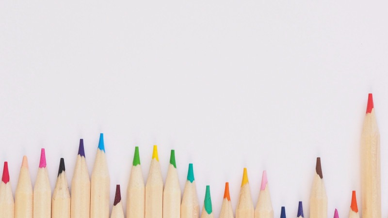 A row of colored pencils arranged in a gradient of vibrant colors, resting against a plain white background. The pencils are sharpened, showcasing their tips in various shades including red, blue, green, yellow, and pink.
