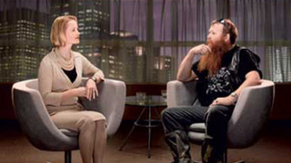 A woman in a beige dress sits in a modern chair, engaged in conversation with a man who has a long beard and wears a black leather vest. The background shows a city skyline through large windows, adding an urban feel to the setting.