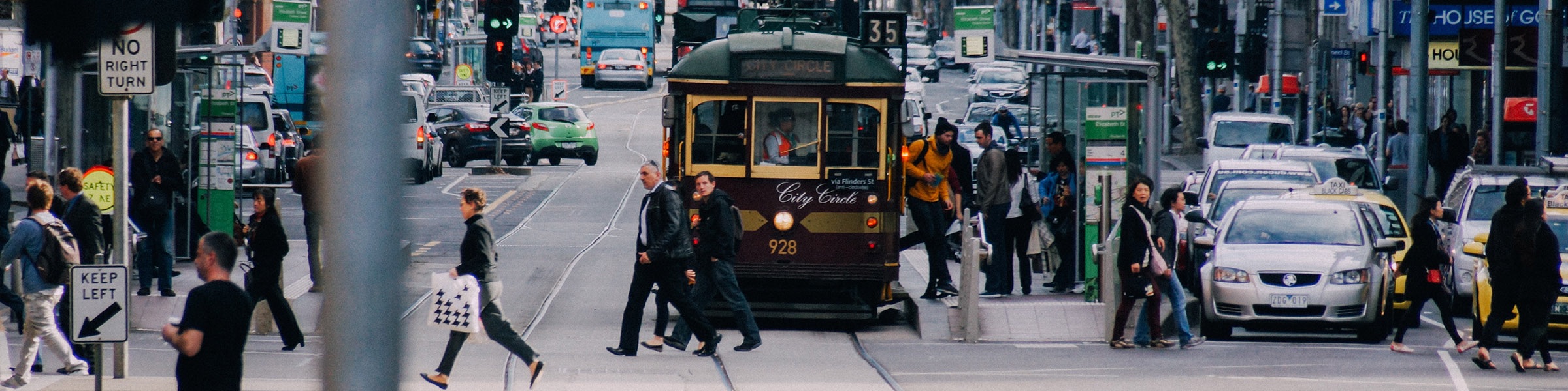 A busy urban street scene features pedestrians crossing in front of a vintage tram while traffic flows in the background. People are seen walking with bags, waiting at a bus stop, and navigating the crosswalks amidst a variety of vehicles, including cars and taxis.